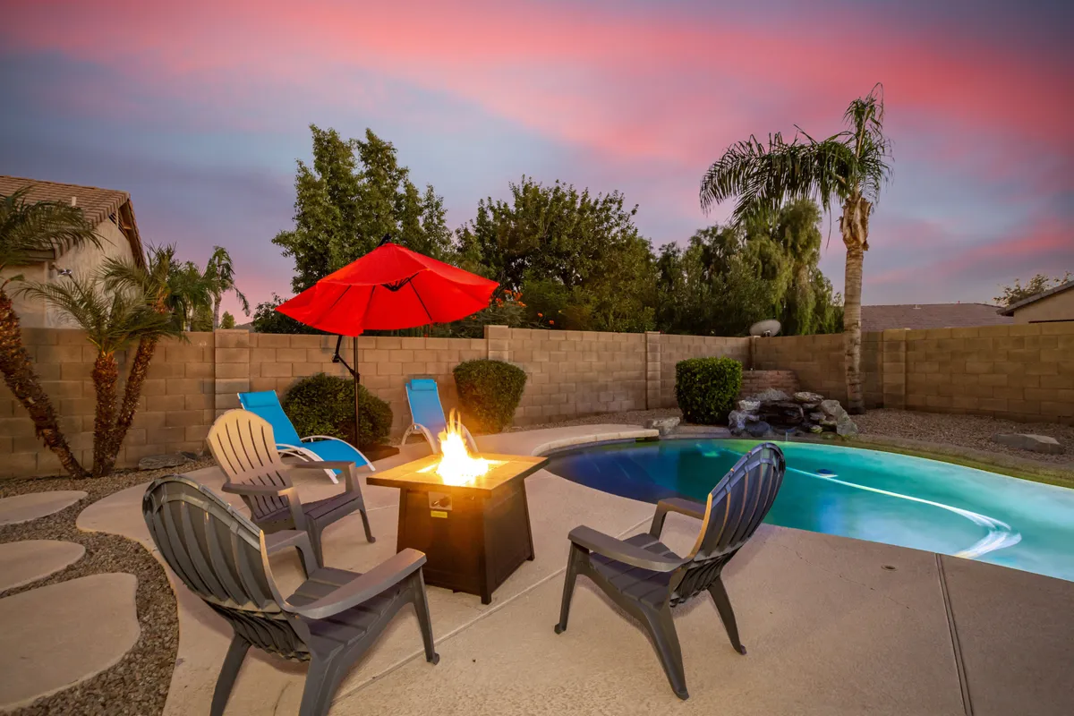 The image centers on a cozy fire pit nestled beside the pool, flanked by lounge chairs and shaded by a red umbrella. The setting sun casts a golden glow, suggesting a seamless transition from pool fun to evening gatherings.