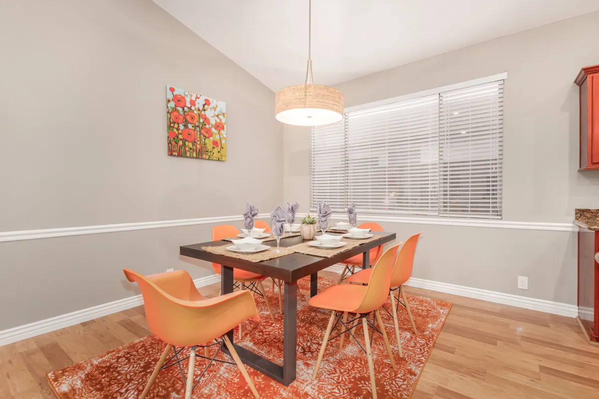 A cheerful dining area with a prominent dining table surrounded by orange chairs, vibrant artwork, and a stylish lighting fixture. The space is inviting for family dinners, celebratory meals, and casual group gatherings indoors.