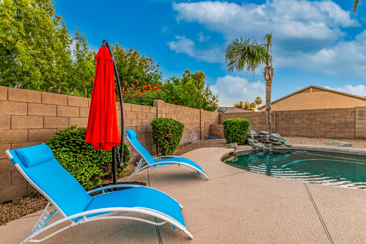 An outdoor view showcases the swimming pool framed by blue lounge chairs and a red umbrella, with green shrubs providing a sense of privacy. The arrangement suggests a space for socializing, sunbathing, and enjoying leisurely afternoons between group activities.