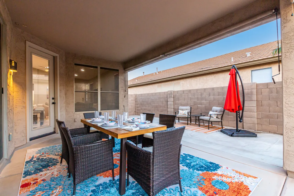 An inviting outdoor dining area features a large table set for eight, woven chairs, a colorful rug, and a hammock chair. Warm lighting and sunset hues evoke a welcoming atmosphere for meals, coffee, and conversation.