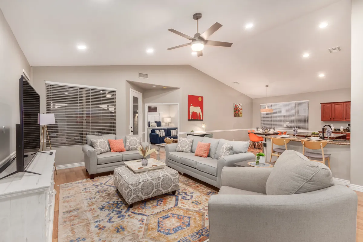 A cozy living room featuring plush sofas, a central coffee table, a large flat-screen TV, and artwork on the walls. The seating is arranged for comfort, conversation, and easy viewing of the TV from multiple spots.