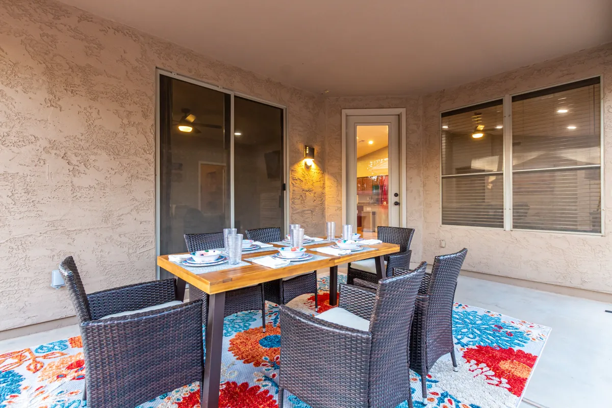 The image displays an inviting outdoor dining area with a table, woven chairs, and a colorful rug, all set under open skies. The scene suggests a vibrant, communal spot for meals, coffee, or casual gatherings in the fresh air.