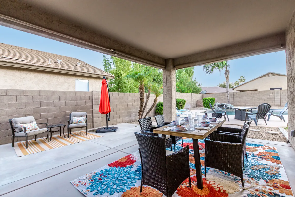 An outdoor dining setup featuring a table with a colorful area rug, red umbrella, and comfortable lounge chairs. The scene is framed by lush greenery and exudes a sense of cozy, casual, and social outdoor living.