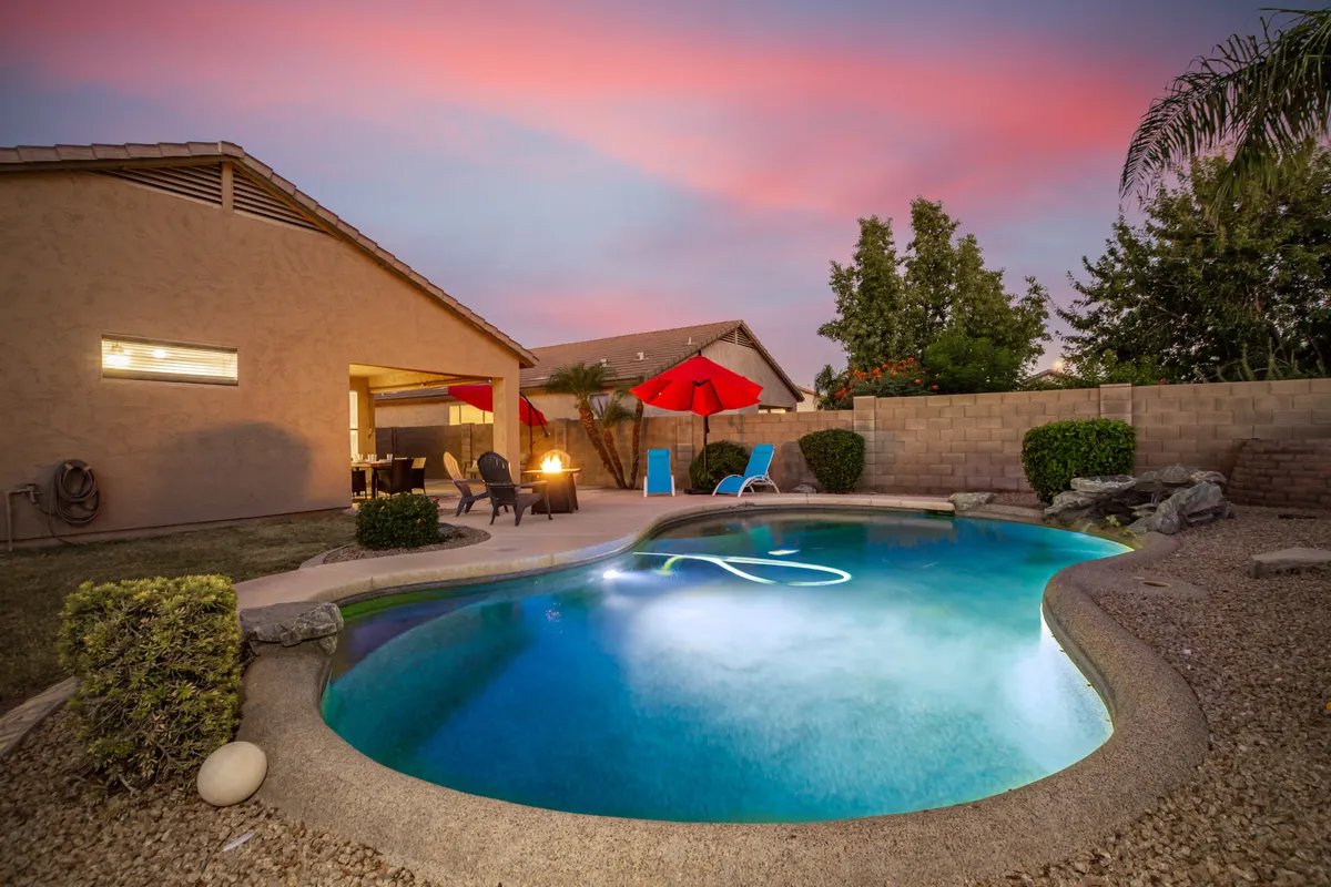 A gorgeous backyard scene featuring a sparkling swimming pool surrounded by plush patio furniture, vibrant red umbrellas, and lush landscaping. The tranquil setting is enhanced by the evening light, welcoming families to gather around the pool or lounge with loved ones. The fire pit and comfortable seating invite fun and relaxation after a day of exploring.