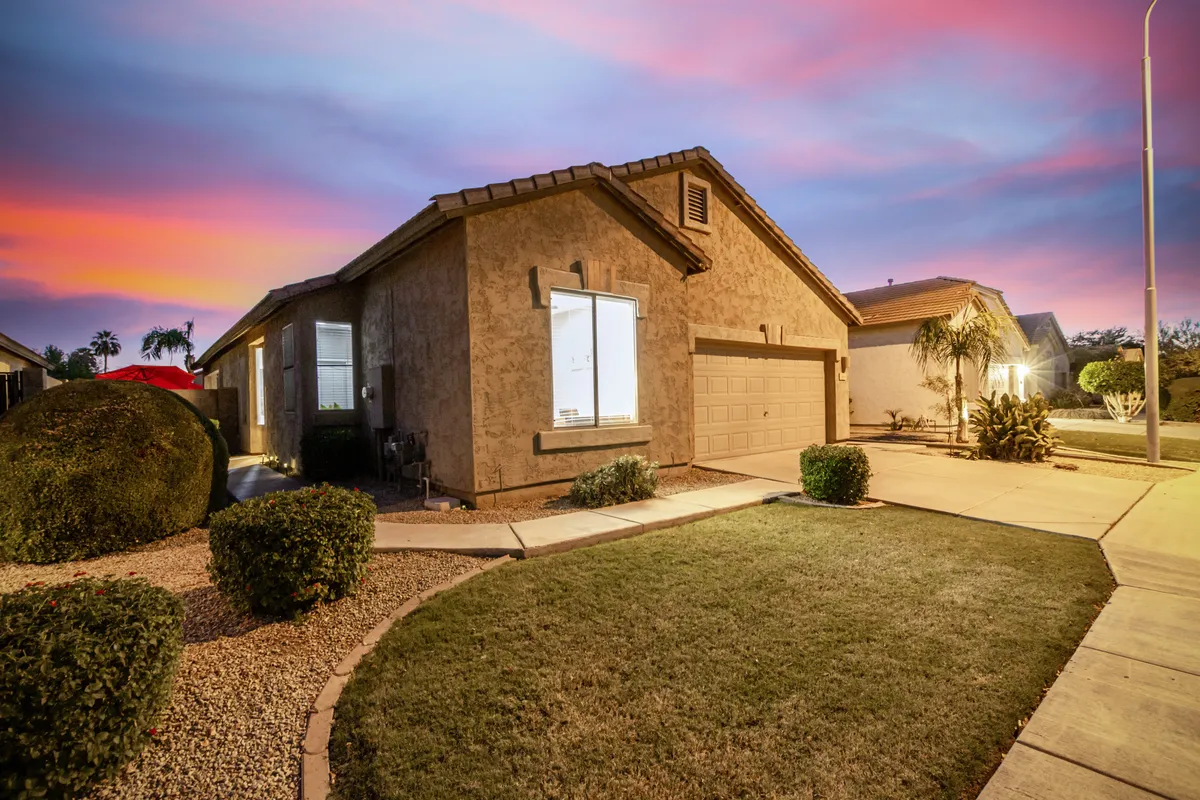 A charming exterior view of the home's front yard at dusk, with an illuminated window casting a warm glow and manicured landscaping enhancing curb appeal. The porch area suggests a welcoming spot to relax after a day exploring Chandler, reinforcing the home's inviting atmosphere.