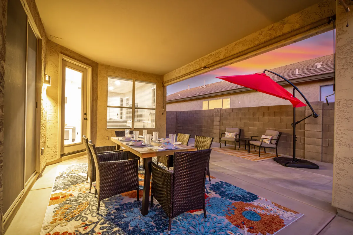 A cozy outdoor dining area featuring a large table with a colorful rug beneath, shaded by a vibrant red umbrella. Surrounding seating offers an inviting space for alfresco meals or evening relaxation, while the view suggests a spacious, private backyard ideal for gatherings.
